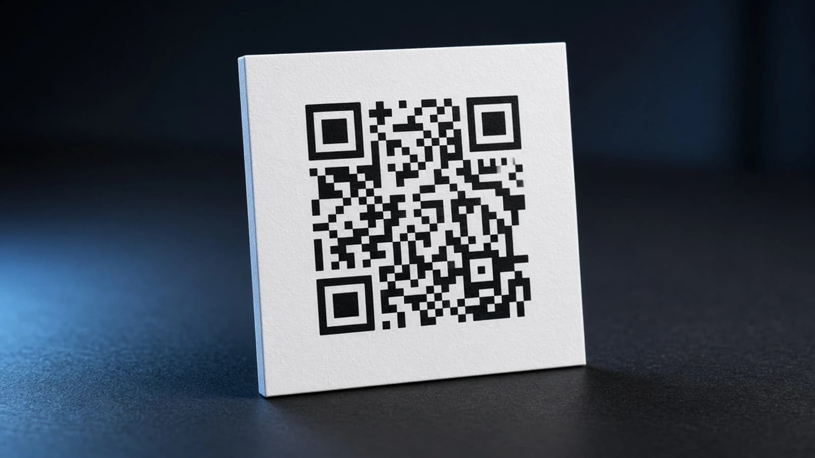 A black-and-white QR code printed on a white card, standing on a dark surface with a blue rim light.